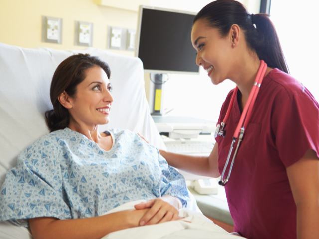 A female patient sits in a hospital bed, while a female nurse in maroon scrubs speaks to her.