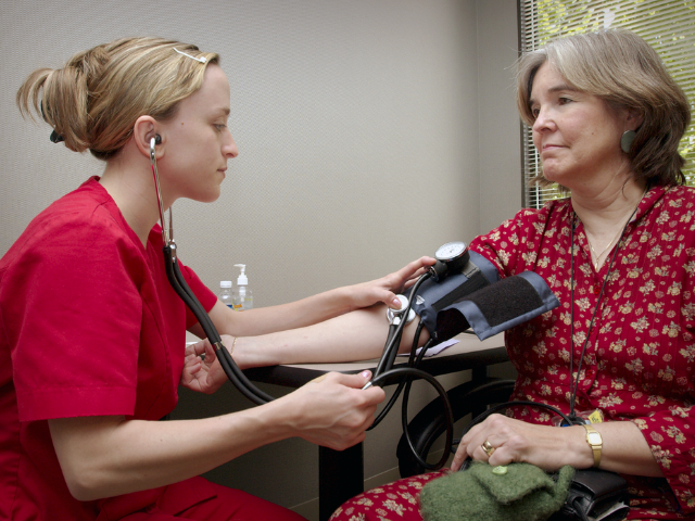 Nurse in red scrubs takes the blood pressure of a woman as part of a public health initiative, similar to what the U.S. surgeon general oversees.